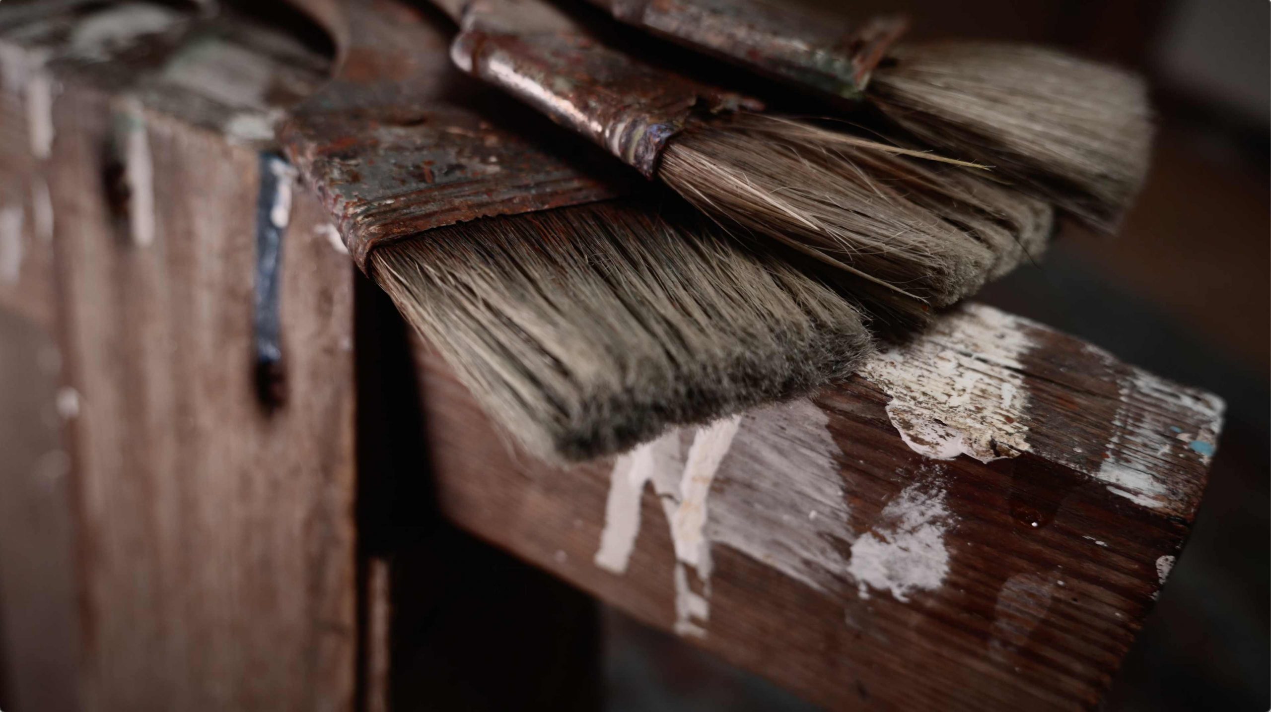 Close-up of three wide, paint-splattered brushes on a rustic wooden stand in the studio of horse and animal painter Hartmut Hellner, with mood lighting highlighting the texture of the bristles.